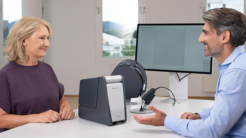 Audiologist explaining hearing aid fitting process to senior couple using ear anatomy model, with Aurical equipment on desk