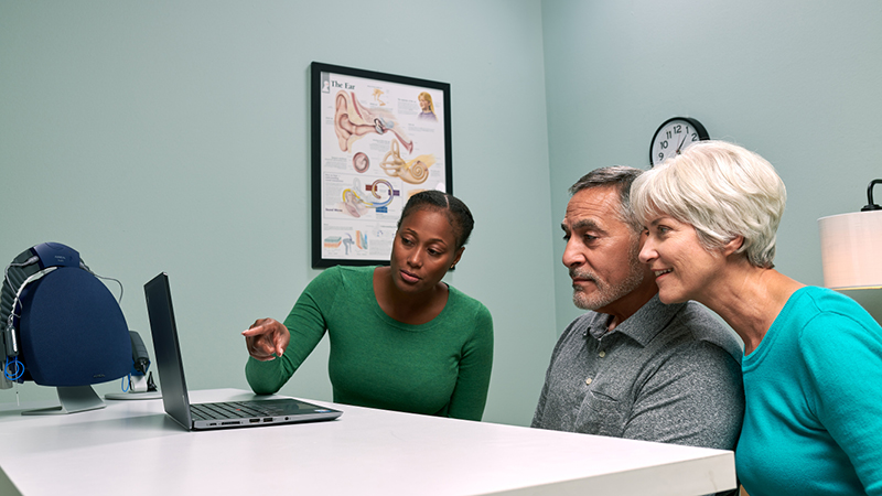 Audiologist explaining hearing aid fitting process to senior couple using ear anatomy model, with Aurical equipment on desk